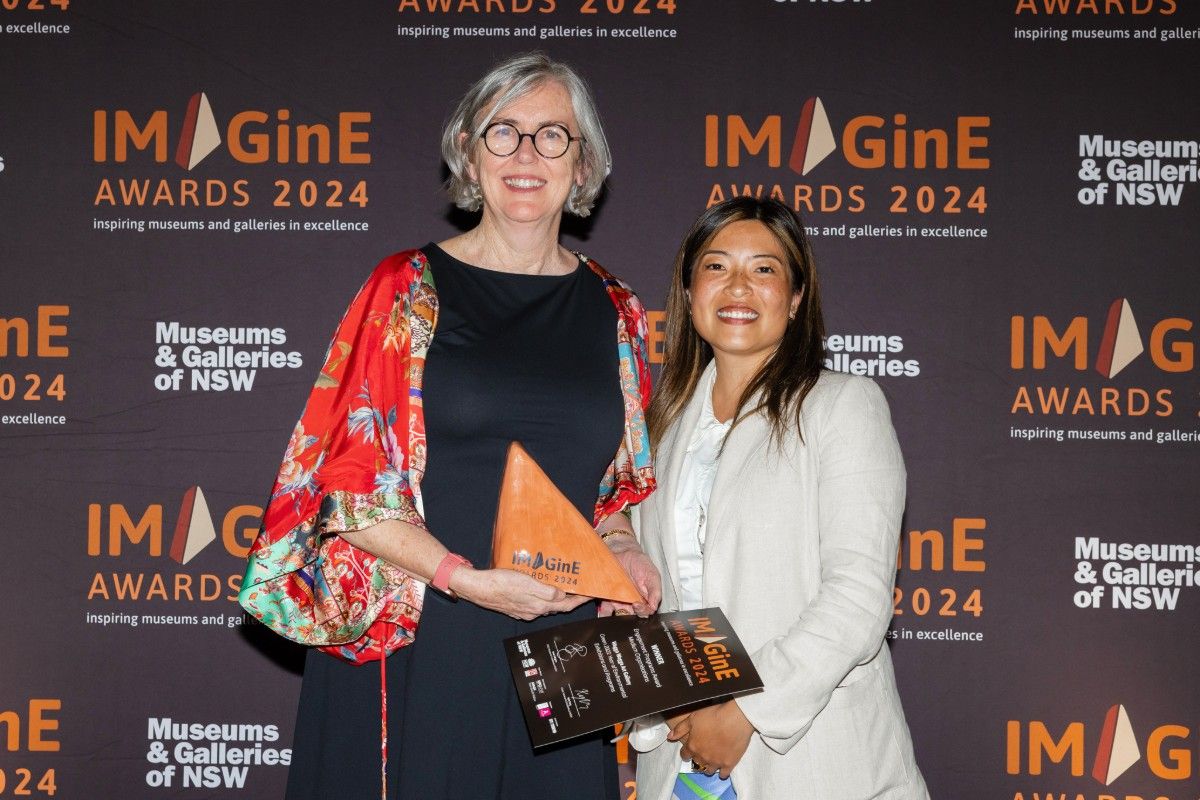 Two women standing on a stage in front of a backdrop sign with IMAgine Awards 2024 printed on it. The woman on the left is holding an award in her hands.