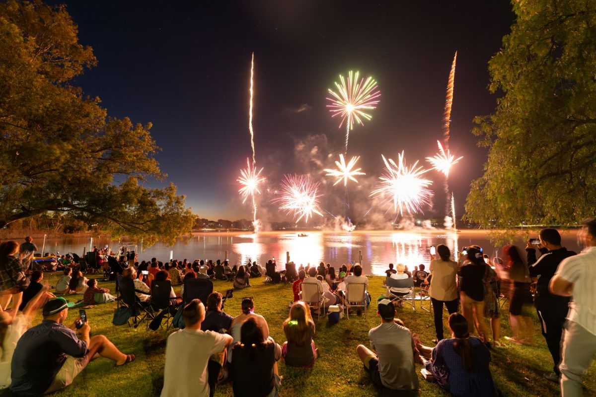 Back view of people and families sitting and standing on grassed foreshores of a lake, watching a fireworks display over the lake.