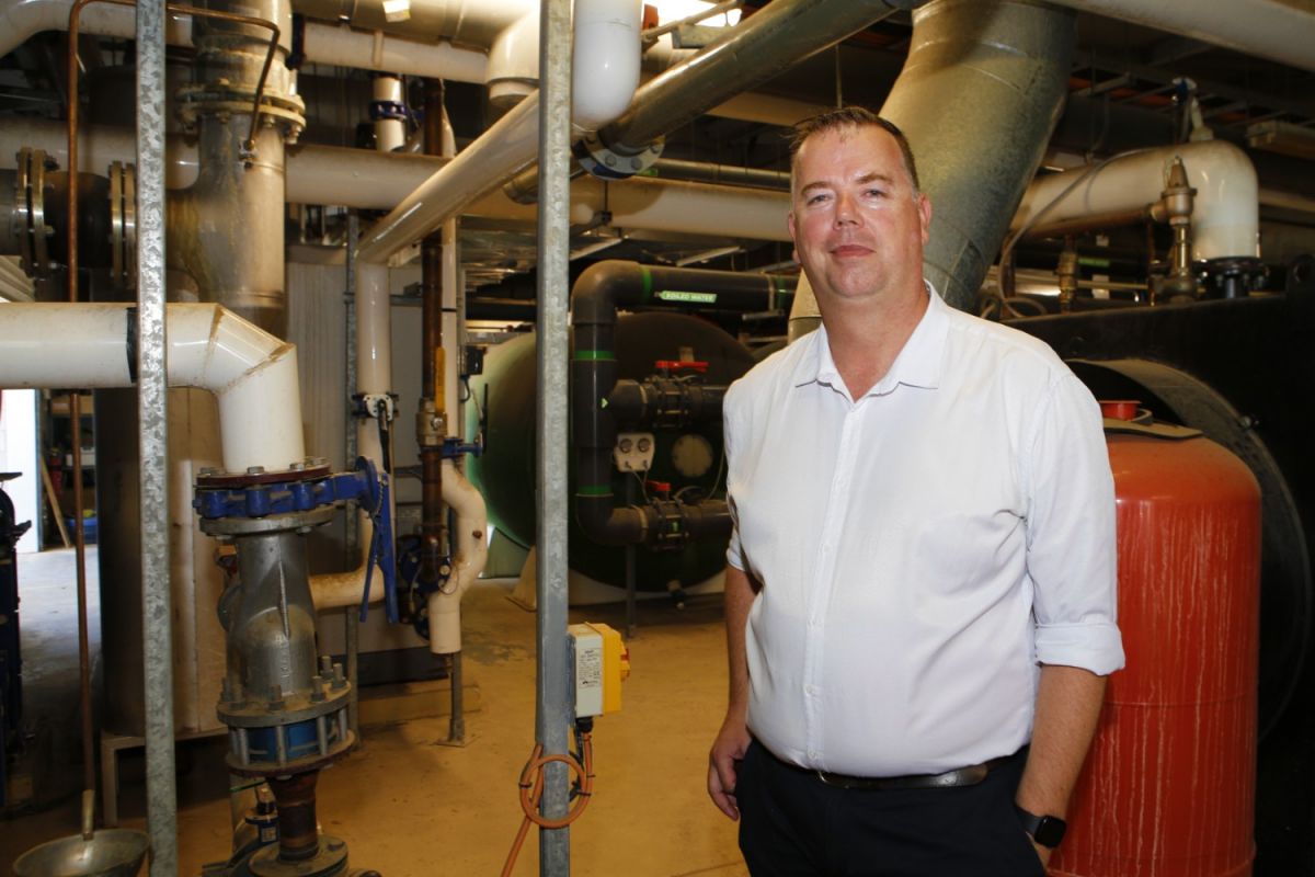 Man in white business shirt standing next to pipes feeding into a gas water heating plant and machinery.