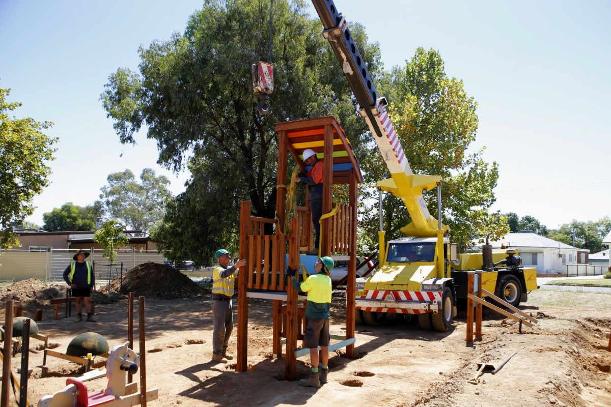 Two men in hi-vis shirts and wearing hard hats hold the frame of a large piece of playground equipment, with a crane and hook in the background.