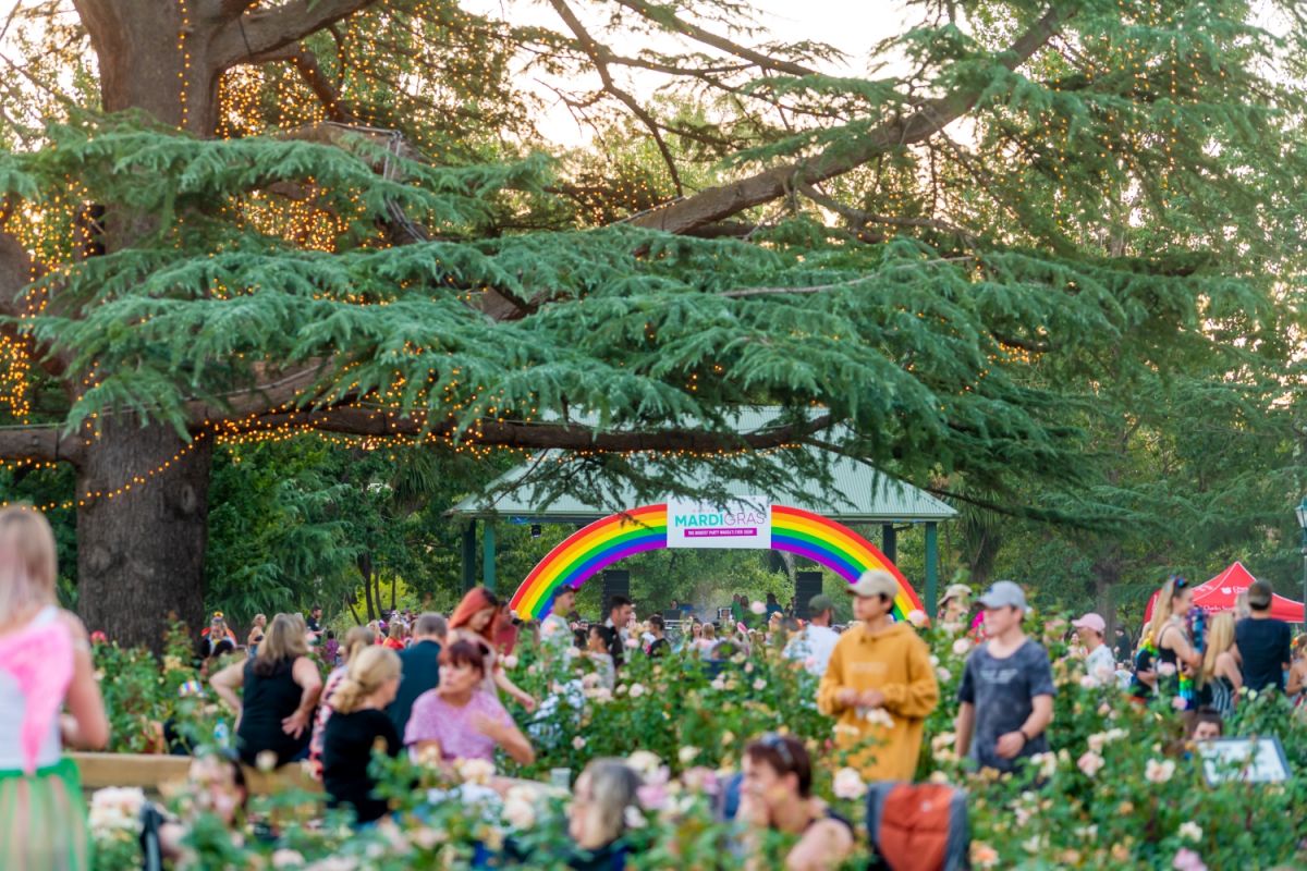 A large crowd of people spread out at the Victory Memorial Gardens, with a large Rainbow displayed in the background.