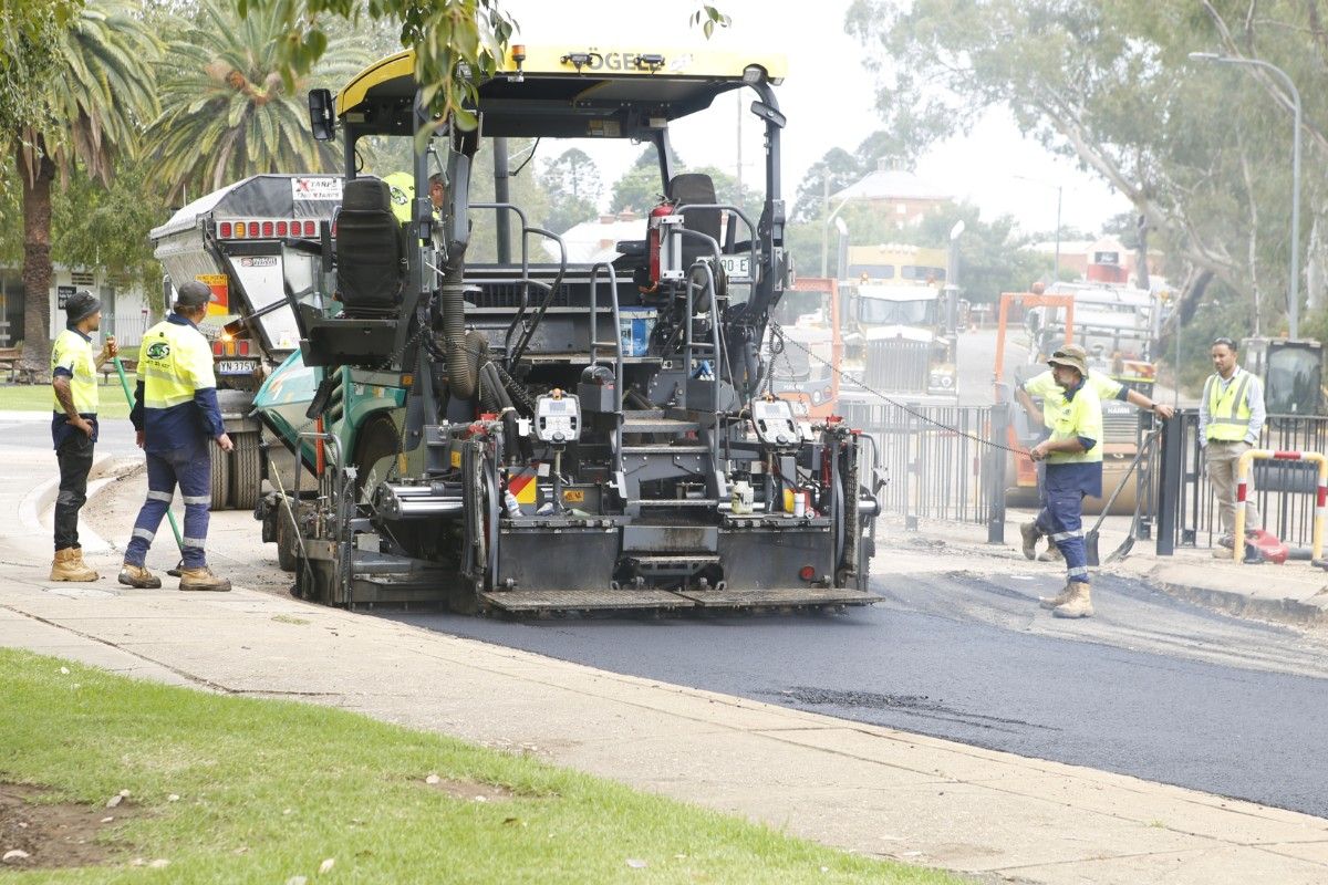 Asphalting machine placing a final layer of deep left asphalt on a road, with road workers with hi-vis vests standing near the machine.