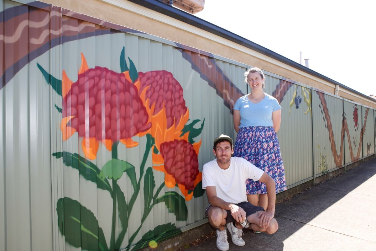 Two people standing outside near a fence painted with a large sunflower mural.