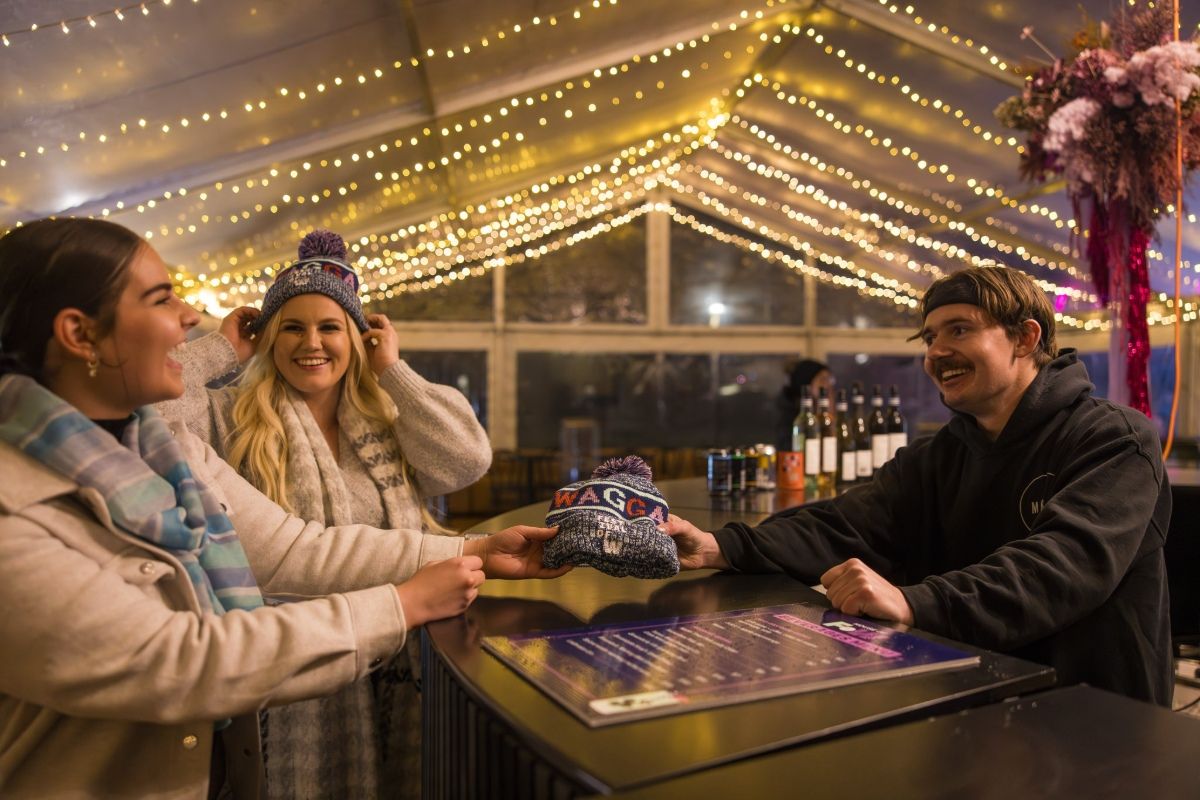 Two women dressed in winter jackets with scarves and a man with a hoodie sit at a table in a marquee with fairy lights on the roof.
