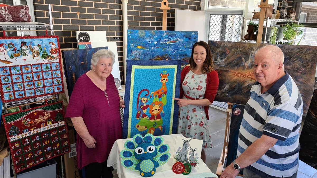 Two women with quilt and one man, all standing in front of paintings