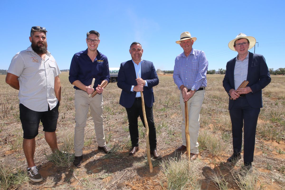 Five men standing side by side in paddock with two holding Aboriginal digging sticks