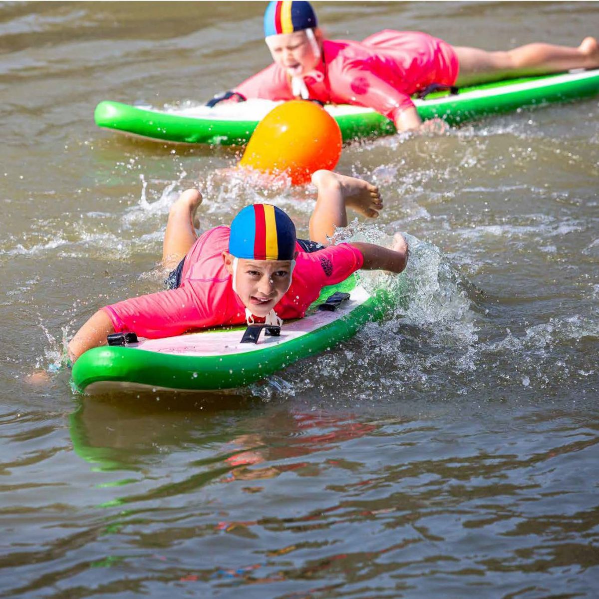 two young boys on safety boards on river, both wearing lifesaver caps