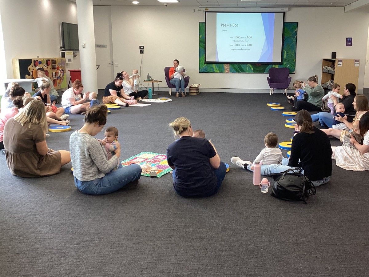 A group of parents and carers sit on the floor in a circle with a baby in their laps.