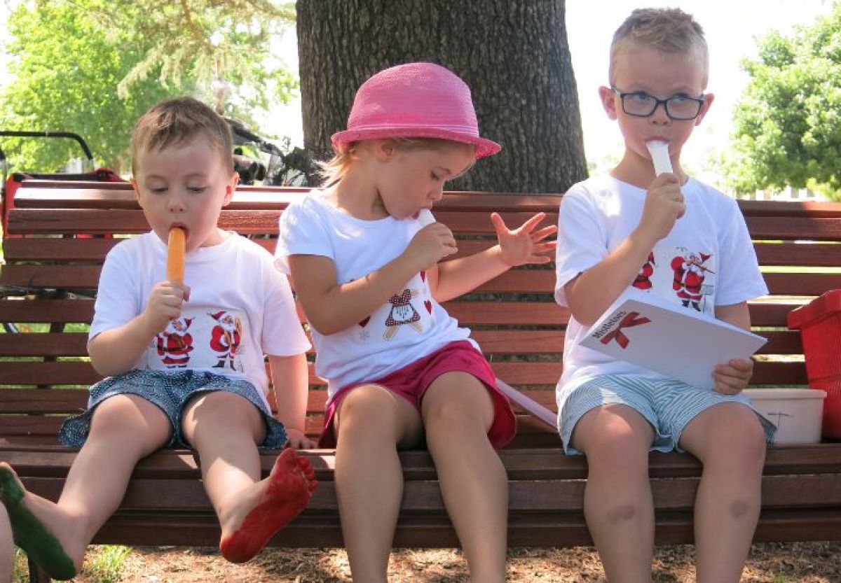 Three young children eating ice blocks on a bench