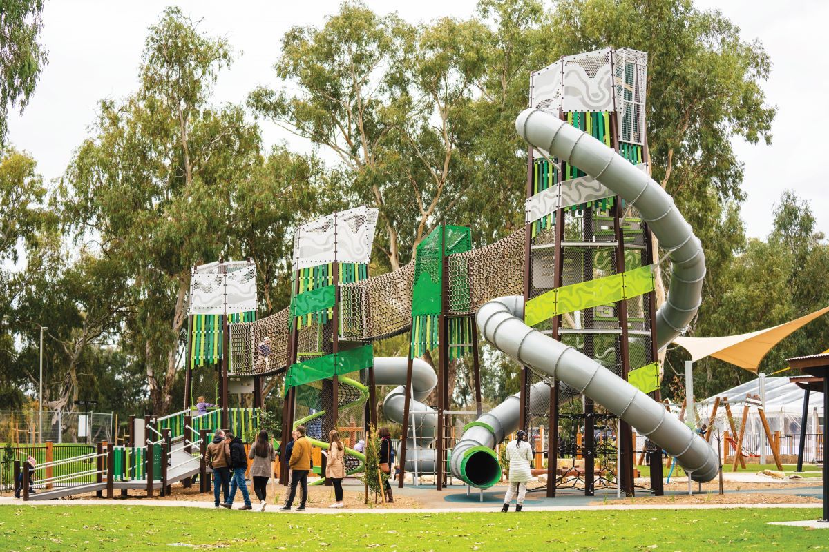 A playground with three large green, brown and silver towers. People are standing in front of the towers. 