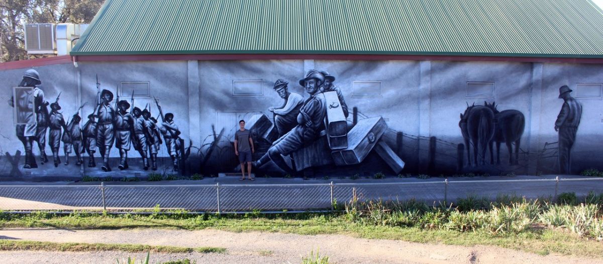 A large black and white mural on an external wall of a community building. The mural depicts soldiers of War.  A young man is standing in front of the mural.