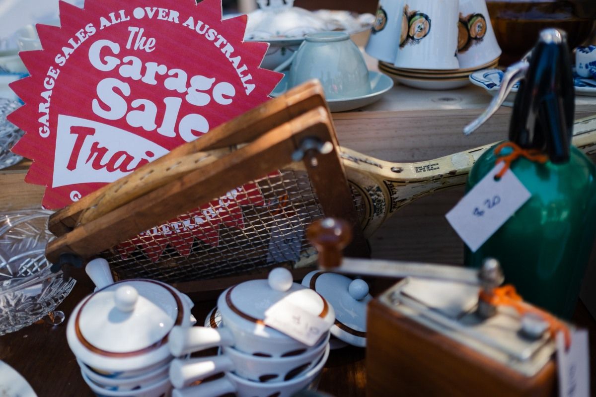 Close up shot of secondhand items for sale on tabletop. The items include retro china, teacups, a tennis racket, and other miscellaneous items. A red circular sign with the text 'The Garage Sale Trail' which sits on the table behind the tennis racket.