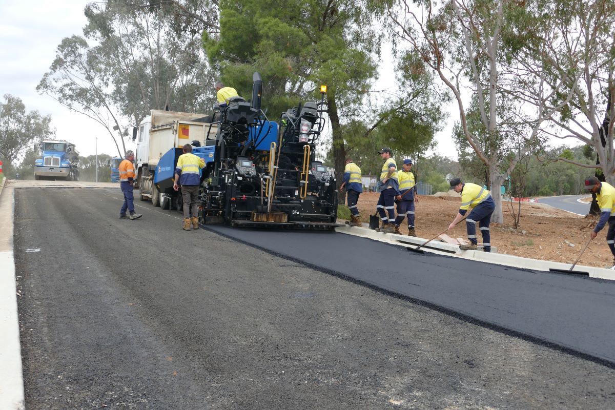 Contractors use a machine and hand tools to apply an asphalt seal to a road surface. 