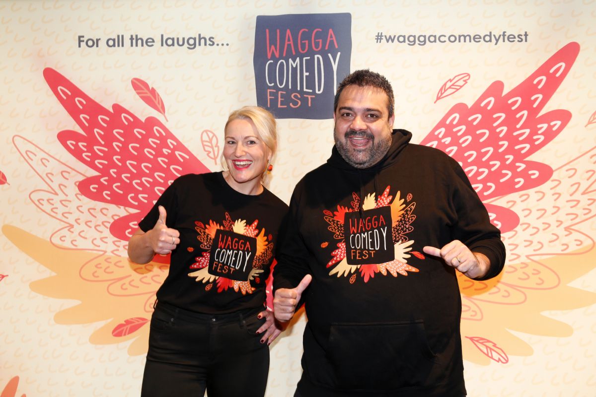 Two people, one female and one male, stand in front of a promotional banner. The pair are smiling and gesticulating, and wear merchandise that promote the upcoming comedy festival.