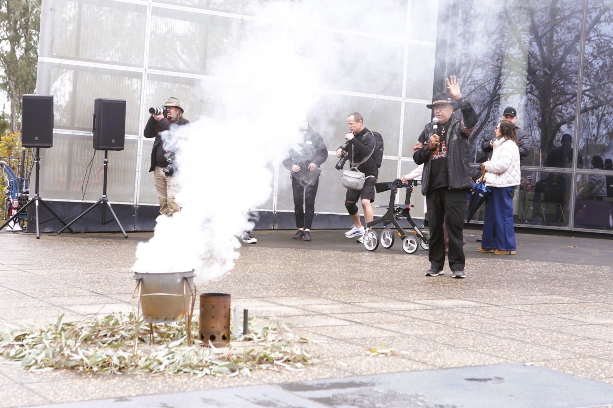 A First Nations Elder addresses the crowd at a Smoking Ceremony, in the foreground is a pale with eucalyptus leaves from which white smoke is rising. 