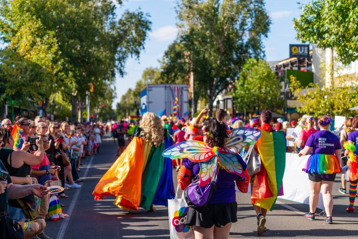 Participants in Wagga Mardi Gras parade, wearing rainbow coloured capes and costumes, walking down main street, with crowd of people on the side lines.