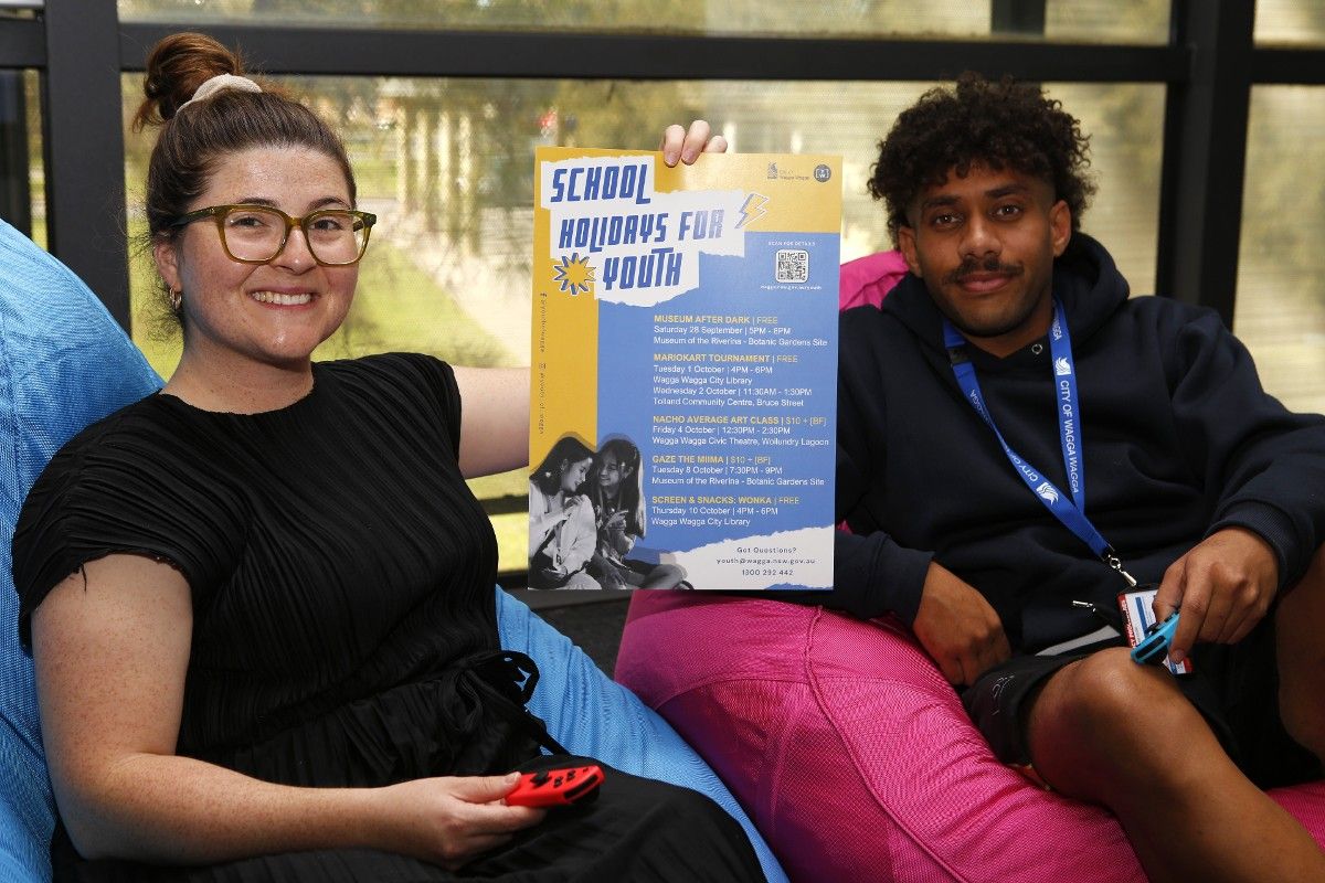 Woman and young man sitting on bean bags, holding Mario Kart controllers in their hands as well as a poster for the School Holidays for Youth program.