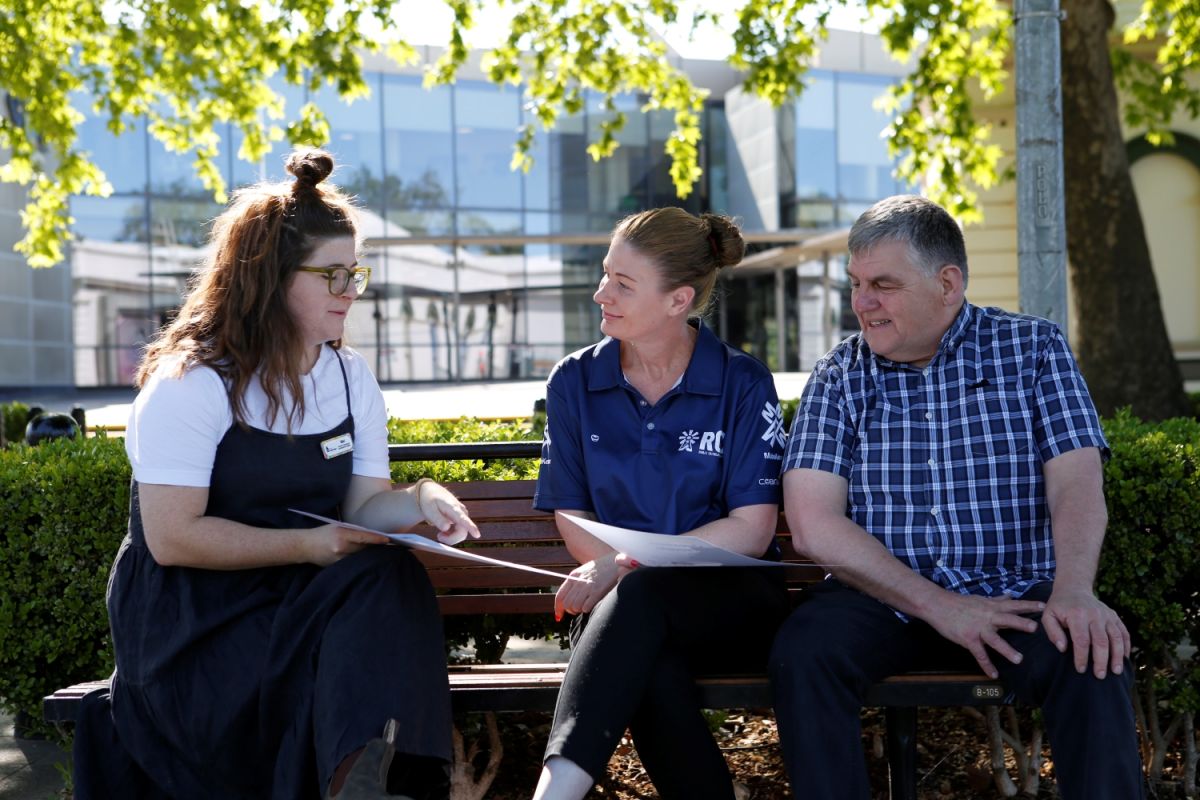 Three people sit on a park bench and gesture to a handheld sign while talking.