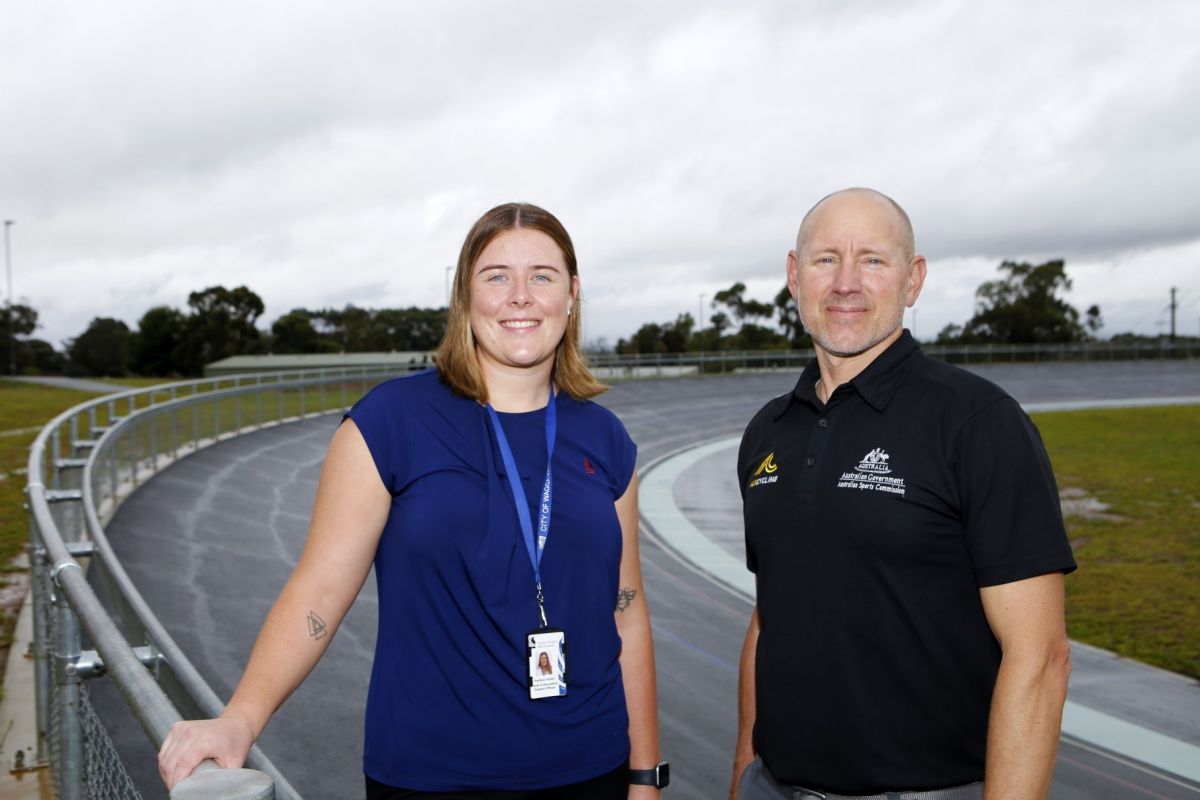 Woman in blue shirt and man in blue shirt standing next to the steel fence around a velodrome track.