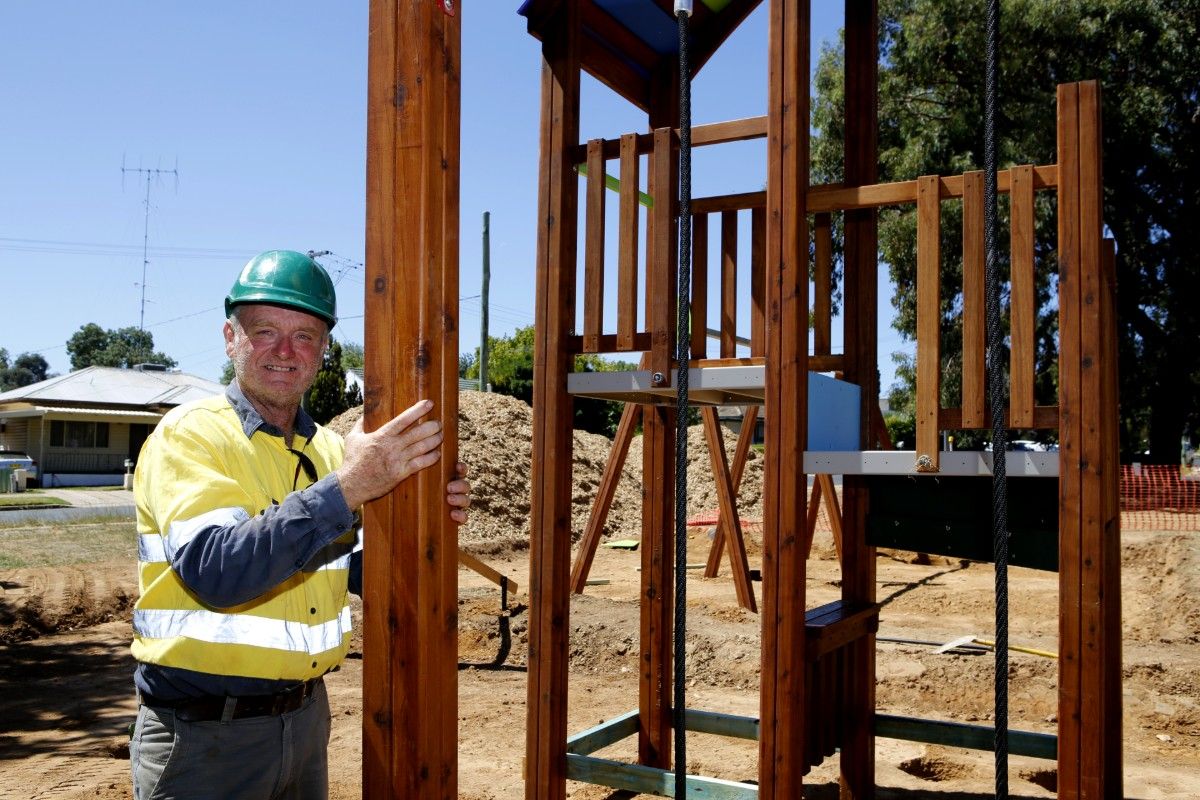 Man in hi-vis shirt and wearing a hard hat holds an upright wooden beam during the installation of new playground equipment.