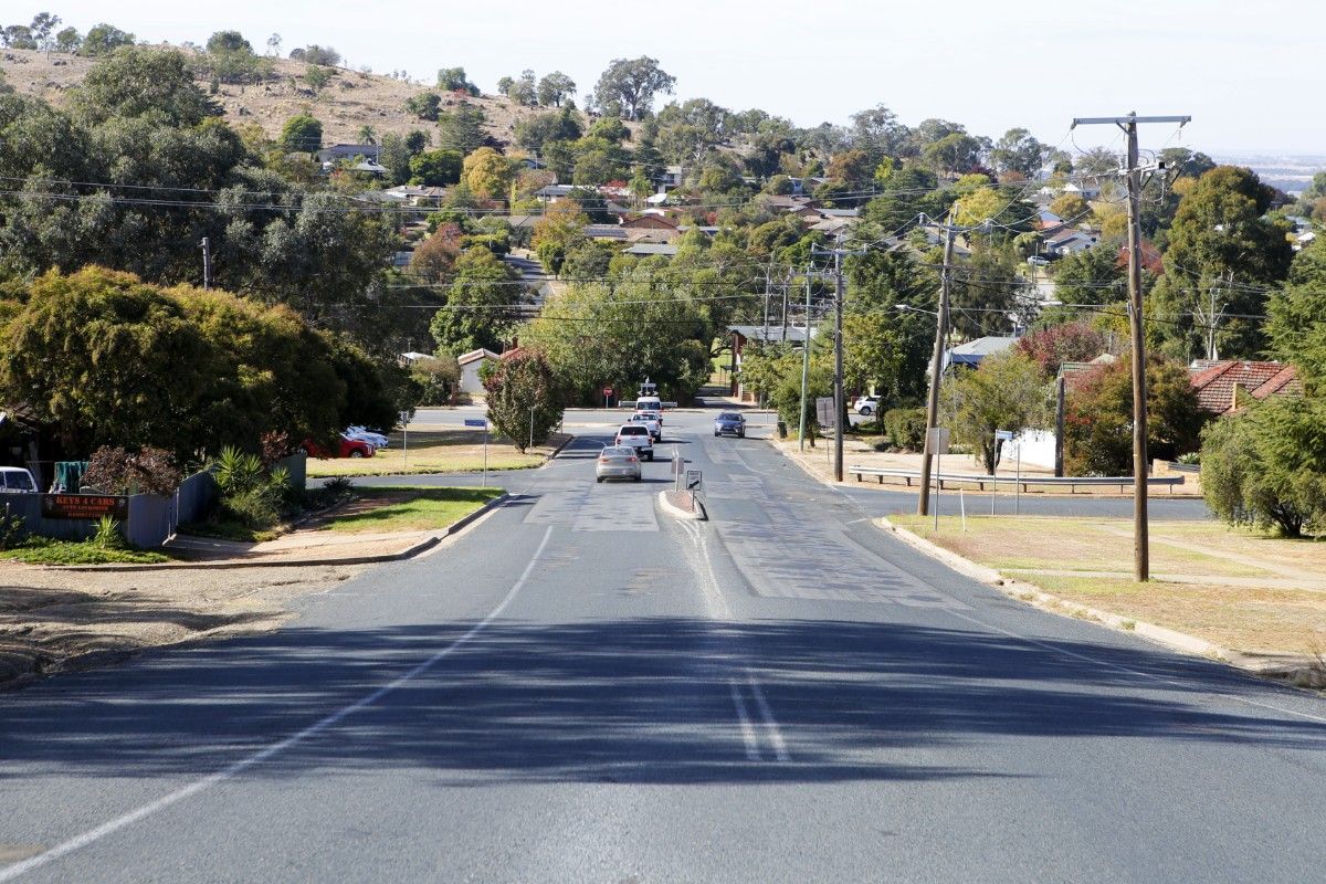 View of a road with patchwork quilt of pothole repairs and cars waiting to run at a t-intersection in the background.