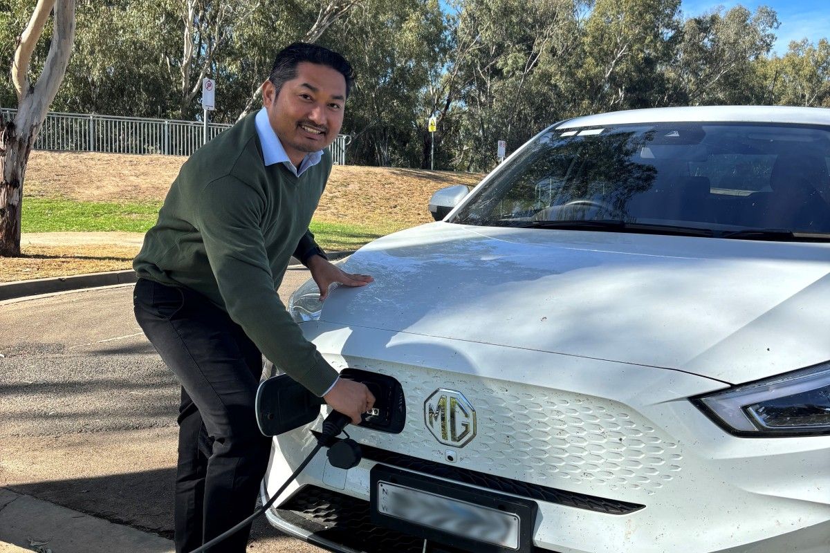 Man wearing a green jumper and black trousers, standing and holding the charging attachment at the front of an electric vehicle in a carpark.