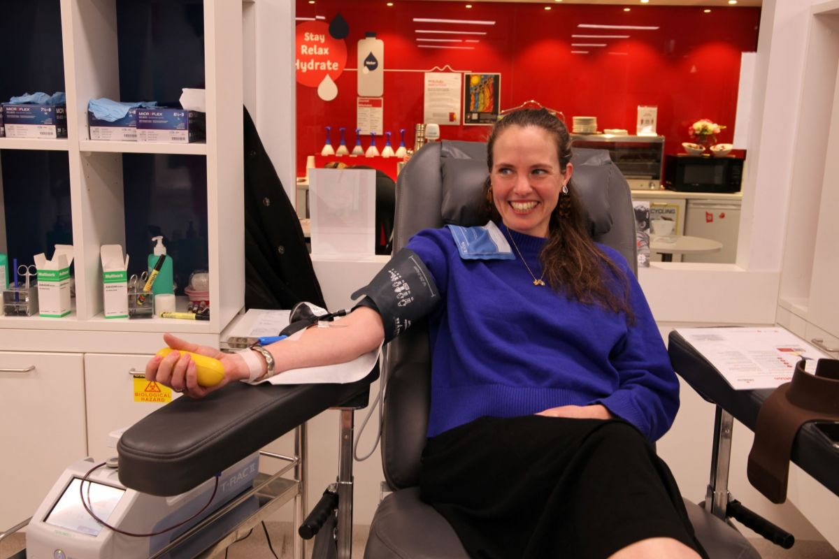 A woman sits in a clinic donating blood. She is wearing a dark blue top and black skirt. She is smiling at someone off camera.