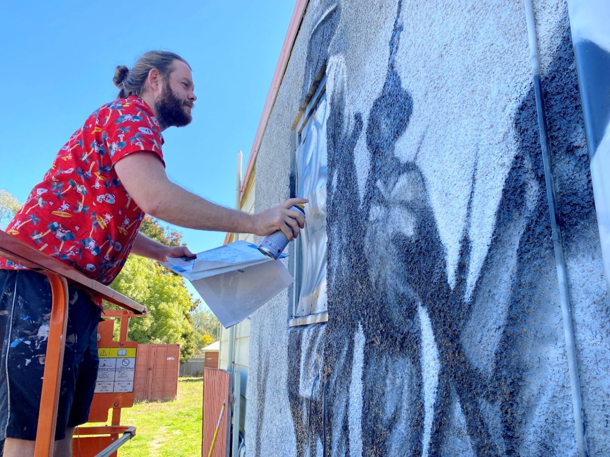 Man leaning over railing of platform and holding a can of spray paint while creating mural on wall 