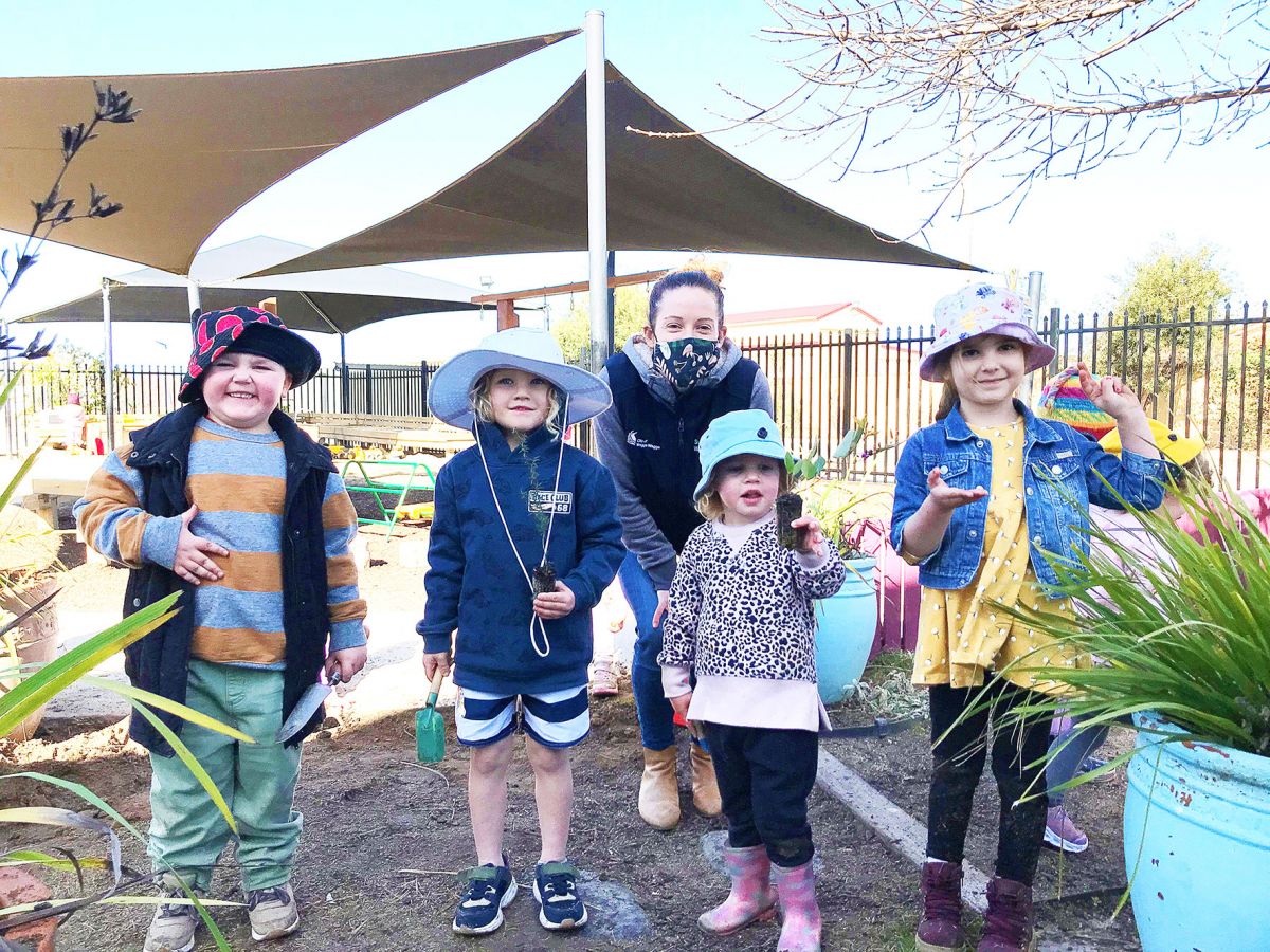 Four young children wearing hats stand in a playground with a young woman.