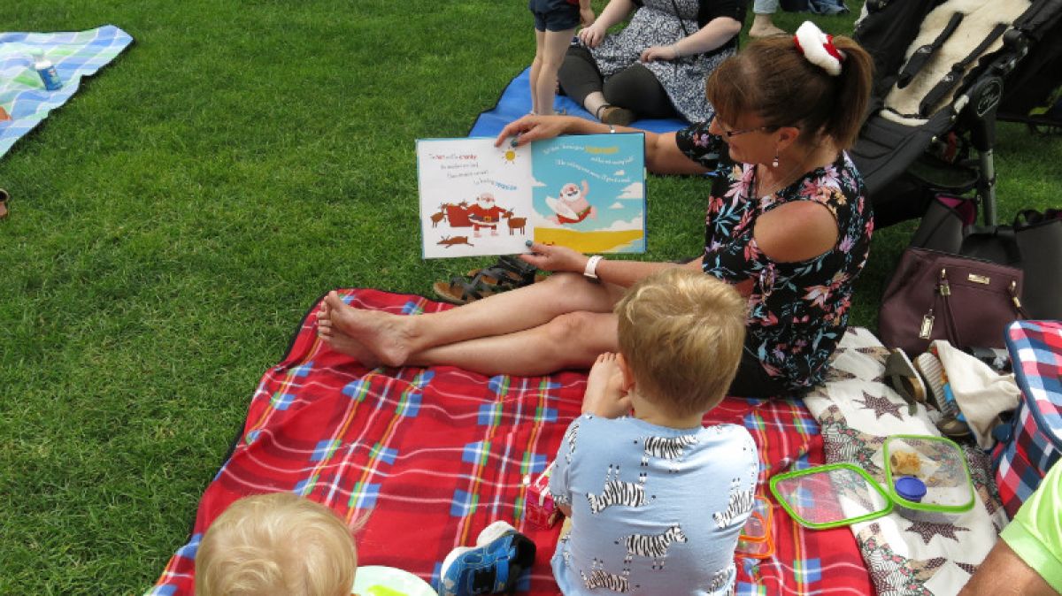 A woman reads a Christmas book to a young child on a picnic rug
