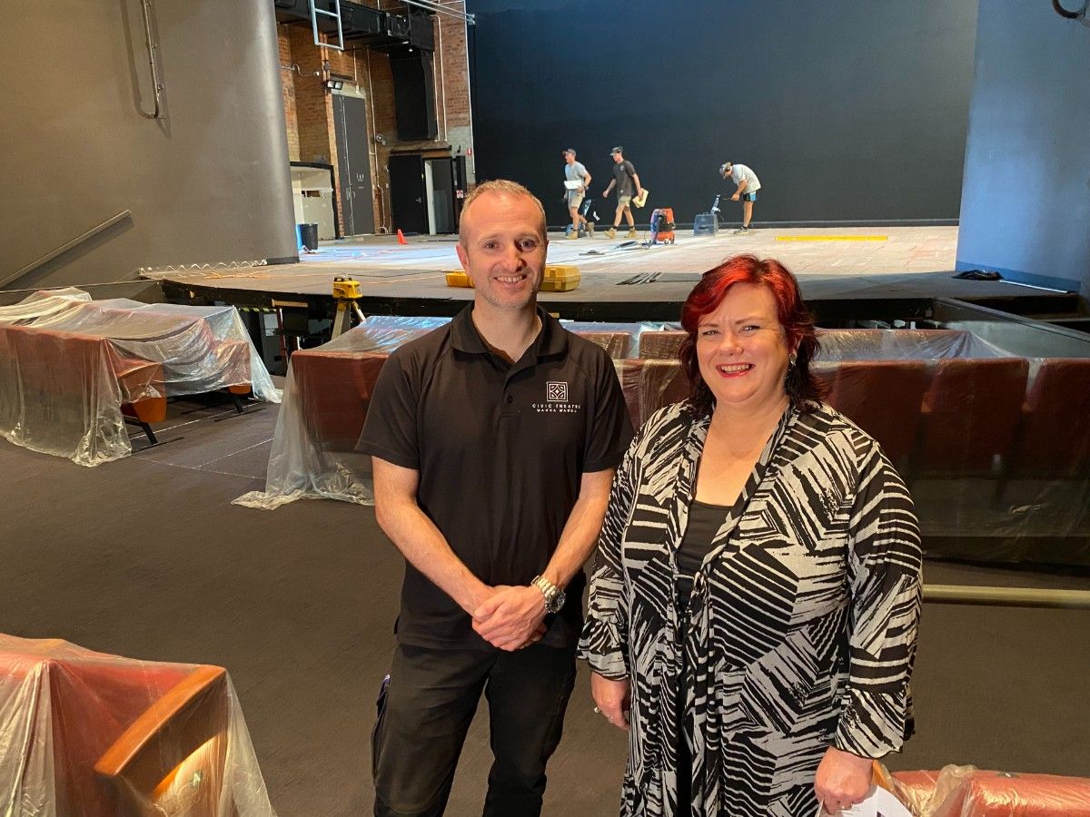 A man and a woman stand in front of a stage. In the background, three contractors are working on the stage.