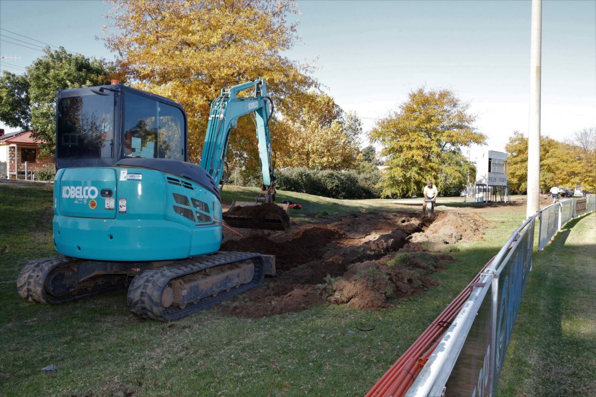 An excavator digs a trench at the side of a sportsground