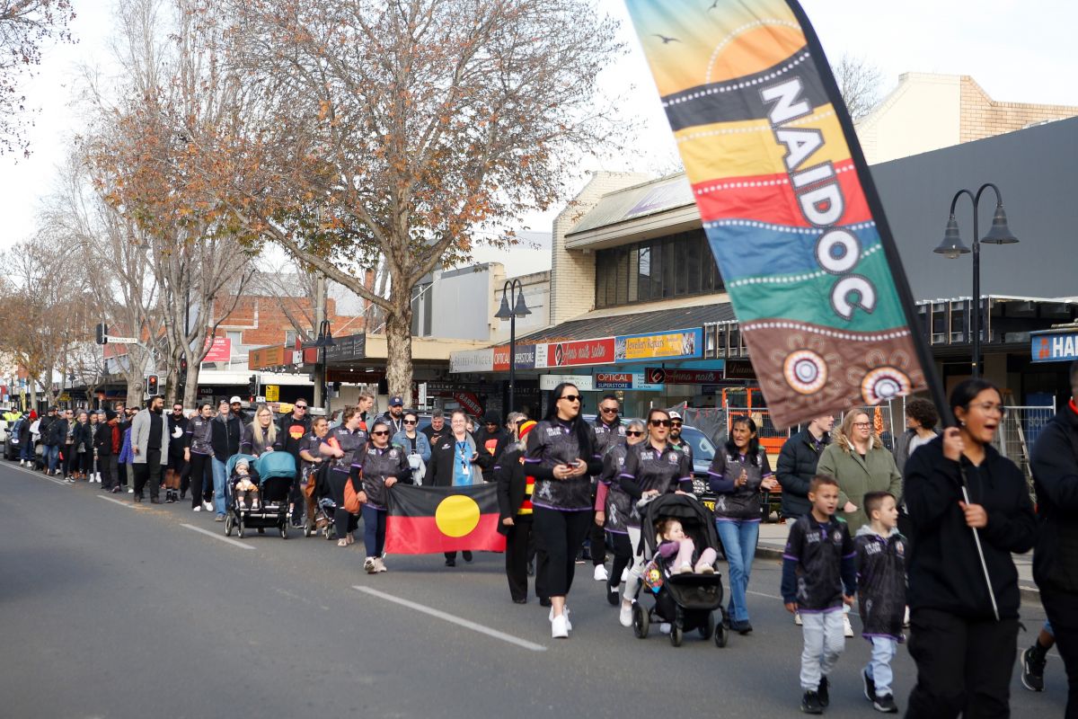 A crowd of people walking down a street past shops holding the Aboriginal flag and banners. 