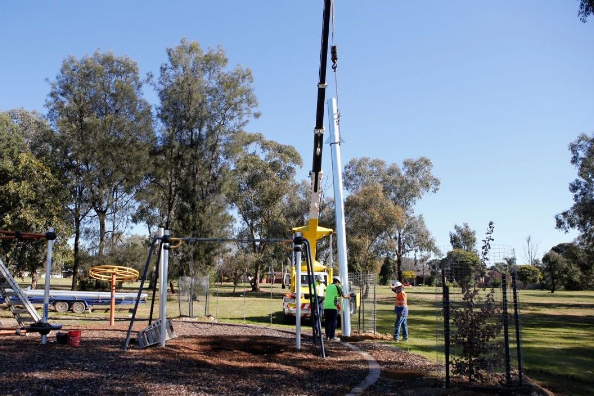 Two men position a large pole in place that is being held up by a crane. To the right is a playground. 