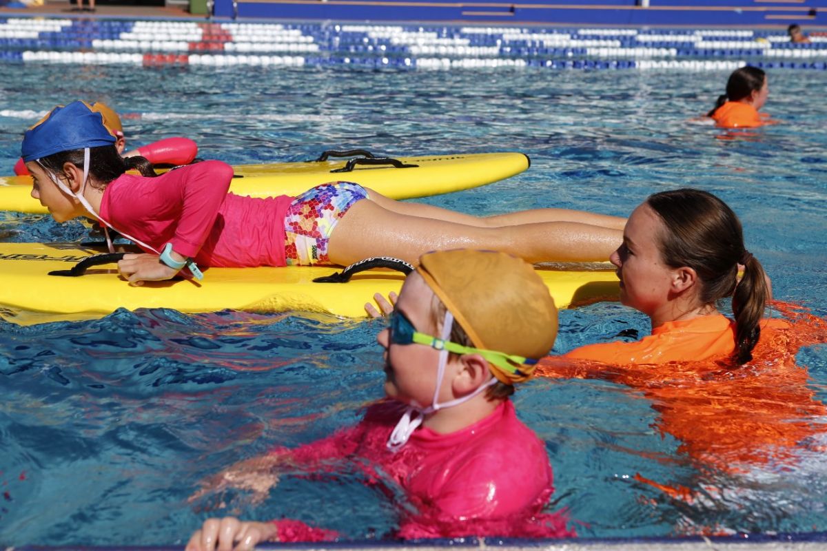 Young woman helps push a young female on a rescue board in a pool, with side on view of another young person treading water nearby