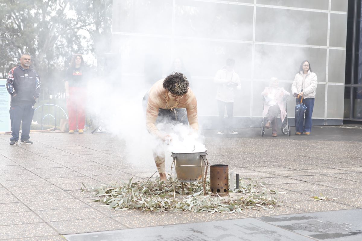 A young First Nations person in traditional dress and wearing ochre on his skin, leans over a pale of eucalyptus leaves from which white smoke is rising, during a Smoking Ceremony. 