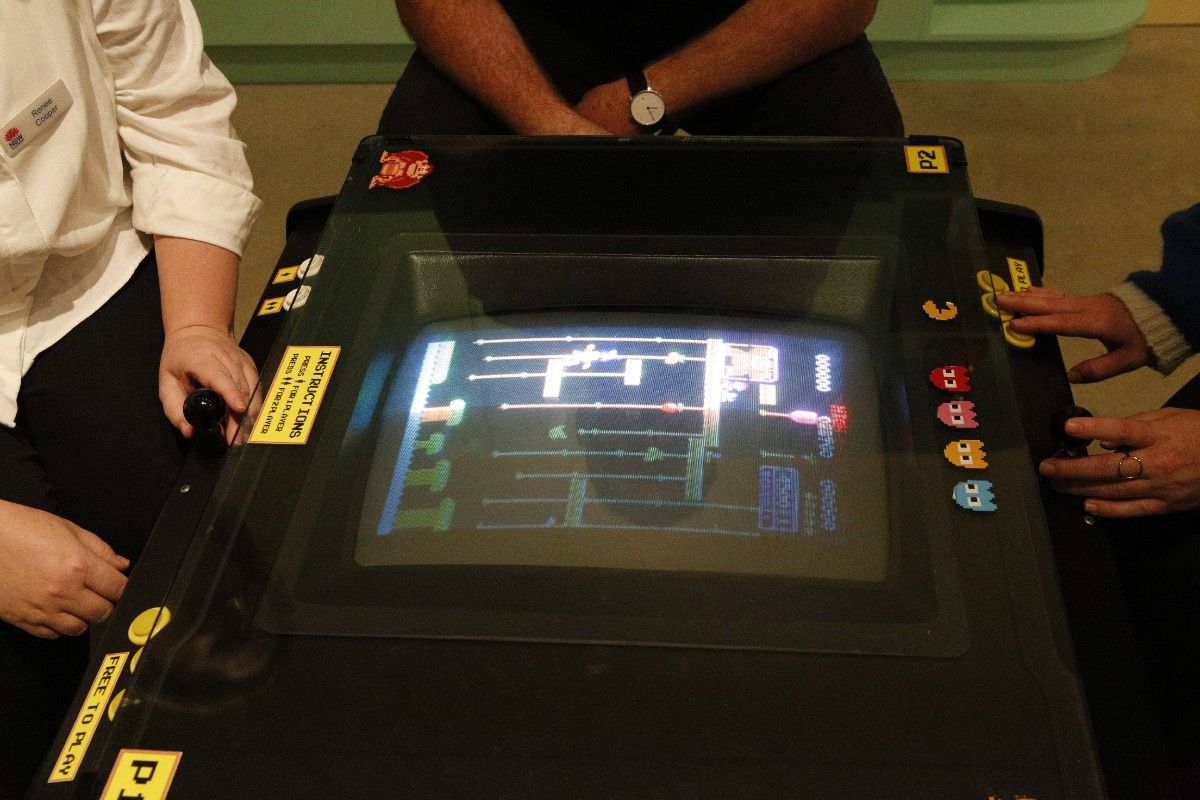 Two women and a man sitting around a retro arcade game machine.