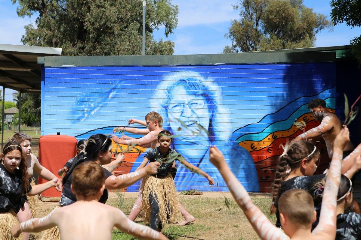 Young First Nations people perform a traditional dance in front of a mural depicting a smiling woman.