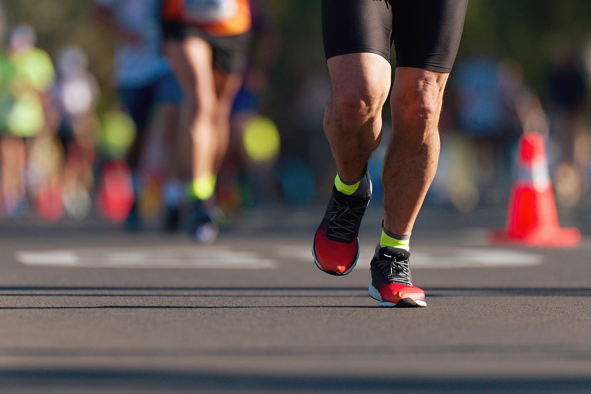 The feet of a person running on a road, behind them, out of focus, is a group of people also running. 