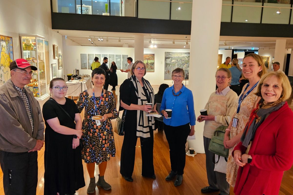 One man and seven women of a range of ages standing in semi-circle, smiling at camera, during a function in an art gallery space.
