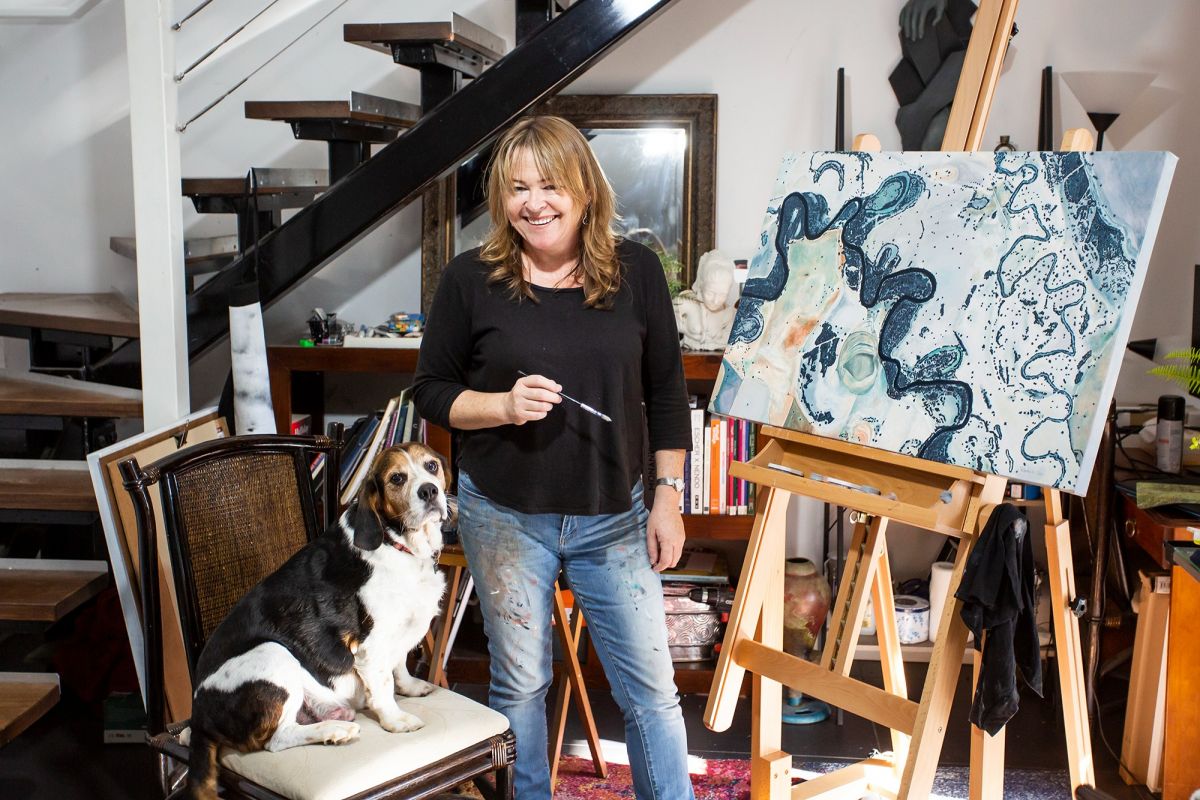 Woman holding a paint brush stands in a room beside an artwork. A small dog sits on a chair beside her.