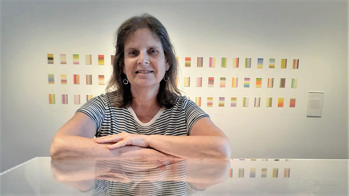 A woman smiles at the camera, she is leaning on a perspex in an art gallery, behind her on the wall are multicoloured cards. 