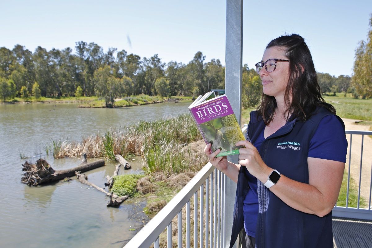 Council Environmental Officer Samantha Pascall stands on the looking platform at Marrambidya Wetlands looking out across the water. She holds a book in her hands titled 'Birds of Australia'.