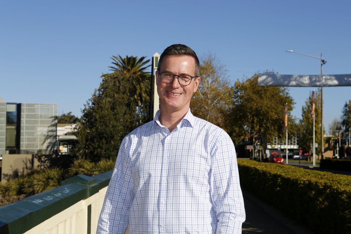 A man in the foreground smiles at he camera. He is wearing business attire and glasses, and stands on a pathway. You can see roads, business fronts, and trees in the background.
