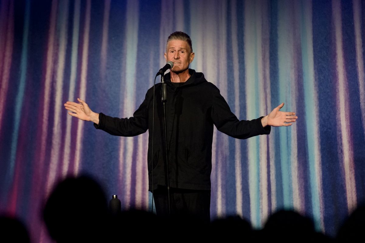 A man dressed in casual clothing speaks into a microphone in front of a theatre curtain. His hands and arms are splayed outwards from his body, palms facing forward. Blurred heads of audience members can be seen.