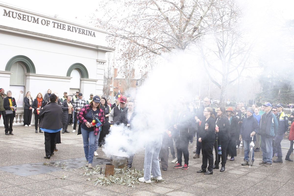 A large group of people gathered in the Civic Centre forecourt, in the centre is a pale of eucalyptus leaves with smoke rising, people are coming forward to 