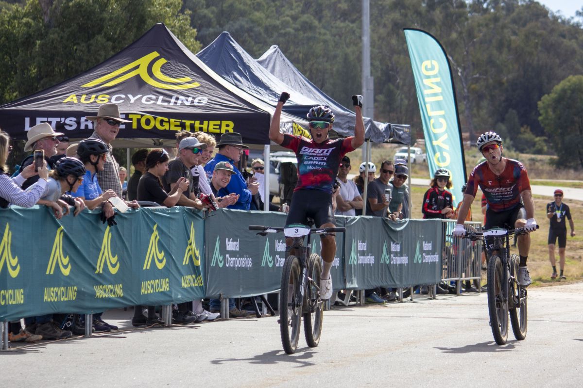 Bike rider with arms in the air, signalling victory as he crosses the finish line ahead of another rider. Crowd lines the track, cheering on the riders.