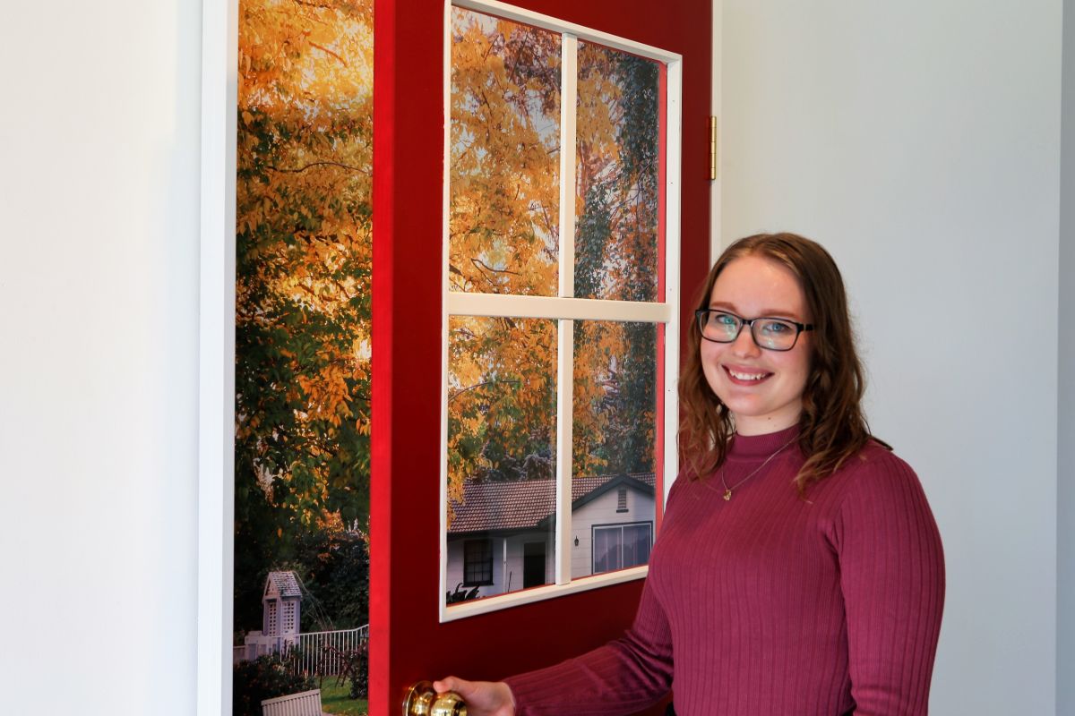 A young woman with light brown hair and glasses holds open a red door that has a large photograph of a house behind it. 