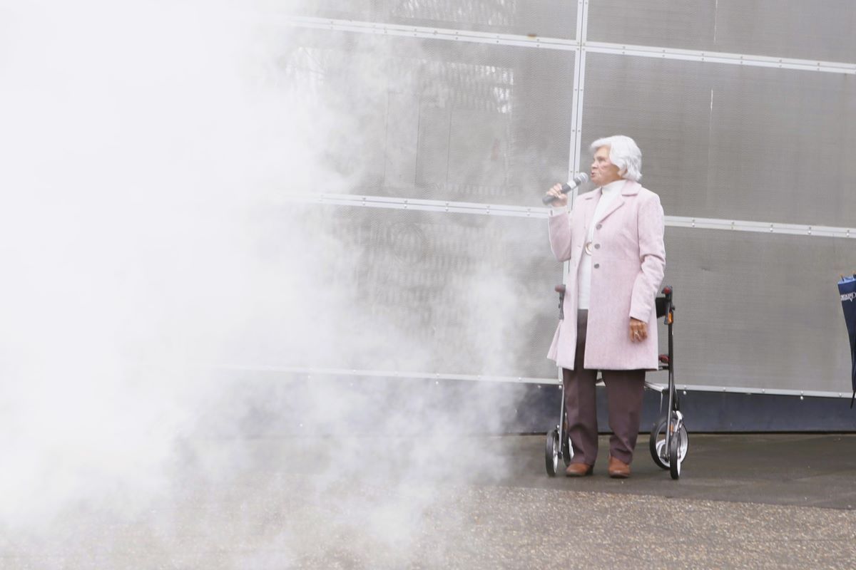 An Elder First Nations woman speaks on a microphone. In the foreground is smoke from a Smoking Ceremony. 