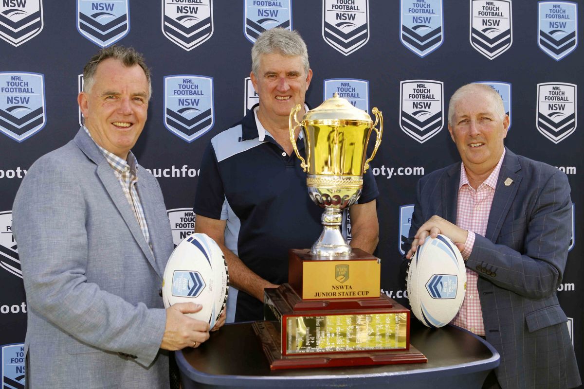 Two men holding touch footballs and third man standing behind trophy on stand in front of NSWTA sign